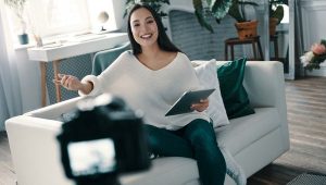 Zu sehen ist eine Frau, die mit einem Tablet auf dem Sofa sitzt und in die Kamera spricht.